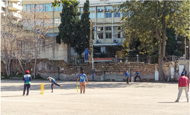 From afar, a ball is thrown at a batter playing cricket with the fielders ready to play defense.