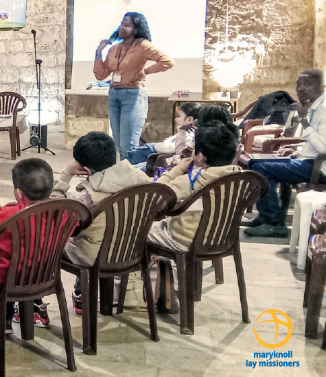 A young adult stands in front of a group of students listening.