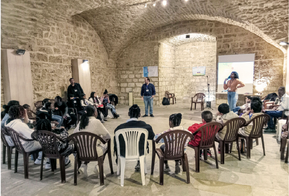 In a large stone room, a circle of children or teenagers sit in plastic chairs listening to three adults leading their gathering.