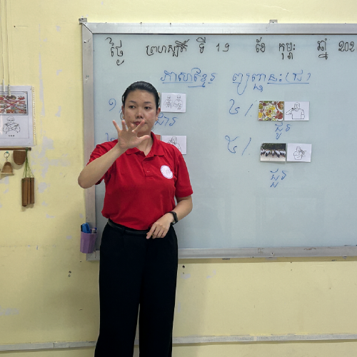 A DDP teacher has her hand out front as she stands in front of a white board with Cambodian words and images.