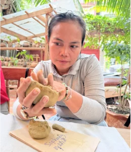 A Cambodian woman forms clay into a bowl while sitting at a table.