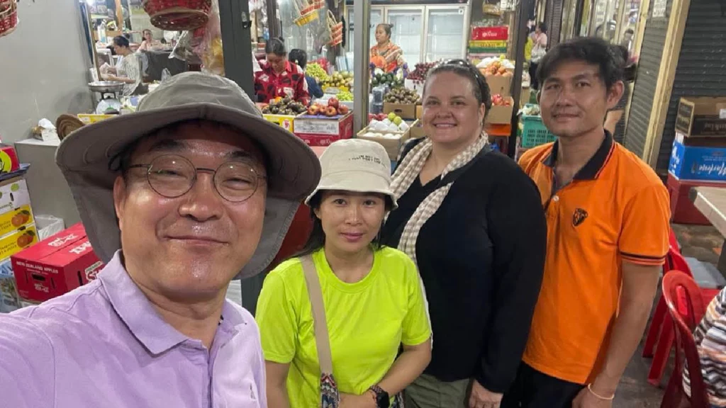 A man in a pinkish shirt and hat, poses with a women in a safety green shirt, Maryknoll Lay Missioner, Julie Lawler wearing a black sweater, and a young man in an orange shirt. They pose in front of a produce market.