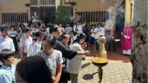 An incense bowl holds several sticks with smoke coming out of them. Maryknoll Lay Missioner, Julie Lawler, adds her own stick of incense into the bowl as a crowd watches on.