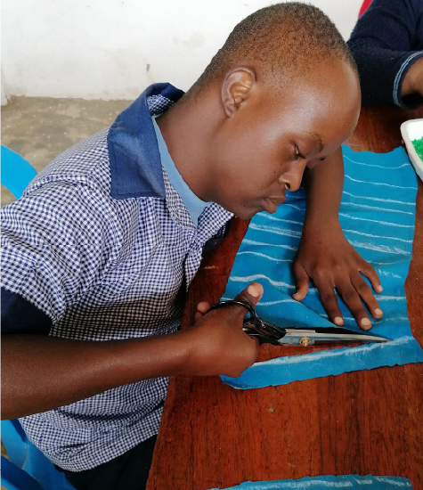 A teenage Tanzanian boy carefully cuts blue fabric.