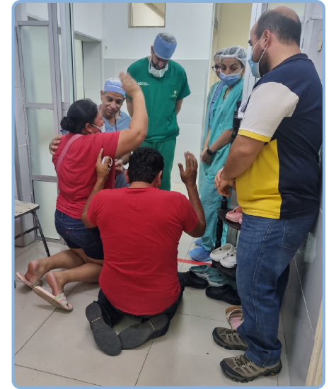 A group of people in a hospital hallway, 2 have their hands up in prayer as 3 doctors bow their heads and Juan Gomez watches.
