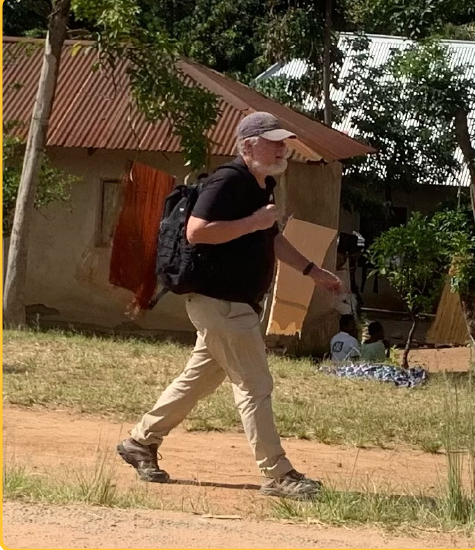 Maryknoll Lay Missioner Mike Lattanzi walks on a dirt road in front of a building and tree.