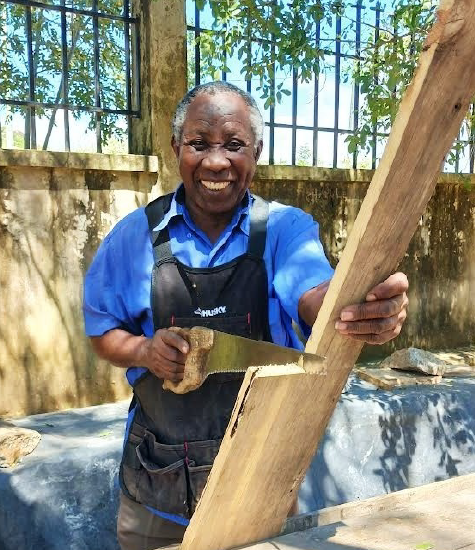 Ernesti the carpenter, shares a smile while holding a saw to a piece of wood.