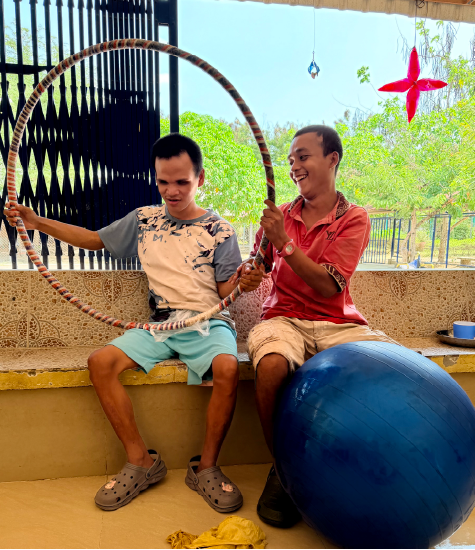 Sitting on a bench, Peu holds up a hula hoop with Bong helping and smiling beside him. A blue yoga ball rests in front of them.
