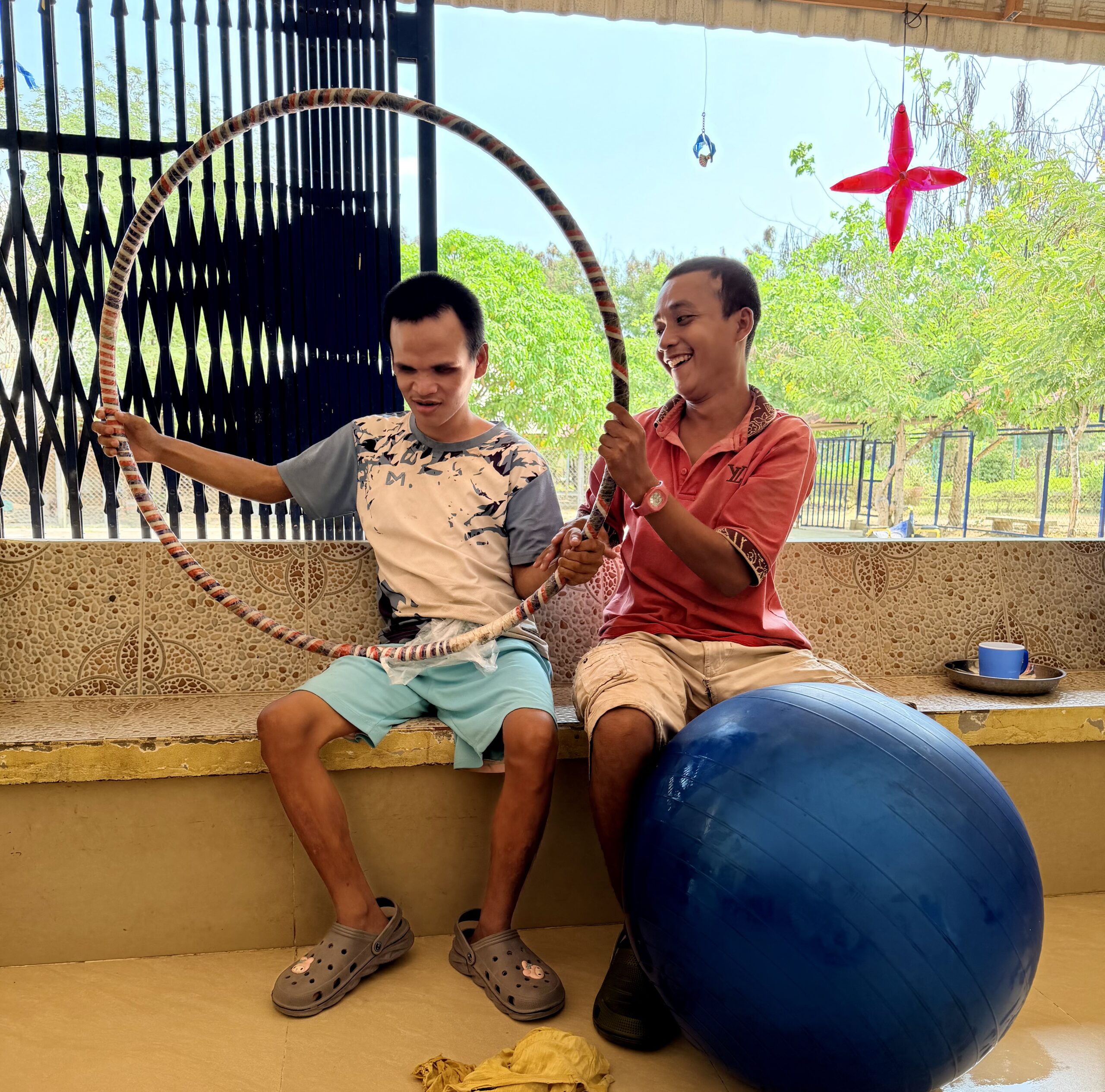 Sitting on a bench, Peu holds up a hula hoop with Bong helping and smiling beside him. A blue yoga ball rests in front of them.